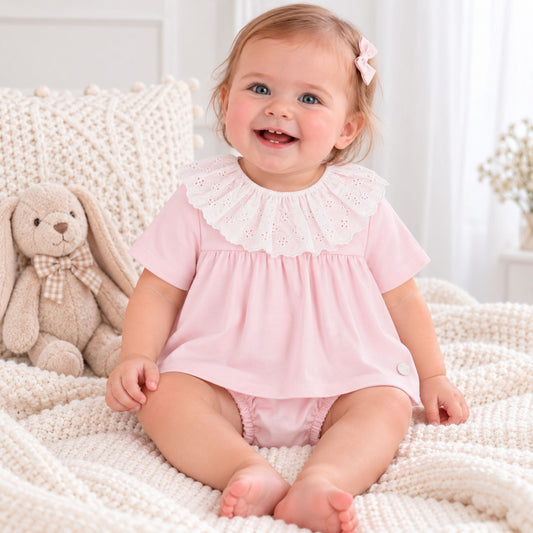 A baby girl sitting on a cream knit blanket next to a plush bunny, wearing a light pink short-sleeve dress with a white eyelet ruffled collar.