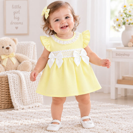 A toddler girl wearing a pale yellow dress with white lace trim, ruffled sleeves, and two large white bows on the chest, standing in a bright nursery-like room with a teddy bear, framed wall art, and a small table in the background.