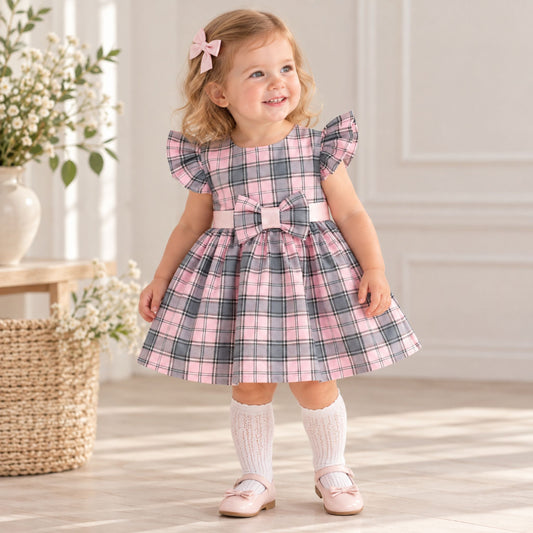 A toddler girl stands indoors wearing a pink and gray plaid dress with flutter sleeves, a matching bow at the waist, white knee-high socks, and pink shoes. The room has soft natural light, a basket, flowers, and a pale paneled wall.
