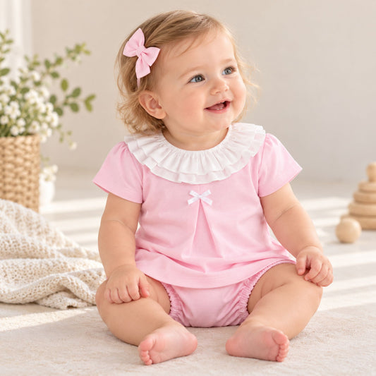 A baby girl sitting on a light beige rug wearing a pink short-sleeve romper with a white ruffled collar and a small white bow at the chest. She has a pink bow in her hair and is smiling in a bright, soft indoor setting.