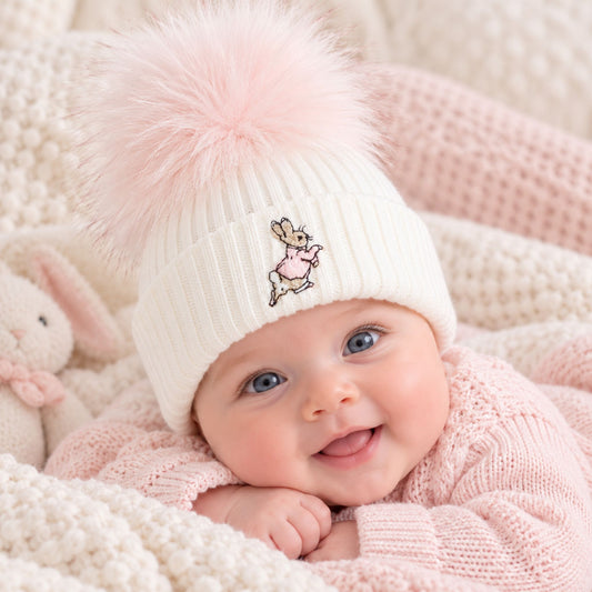 A smiling baby wearing a cream ribbed knit pom-pom beanie with a small embroidered bunny character on the front, lying on a soft pink knit blanket.