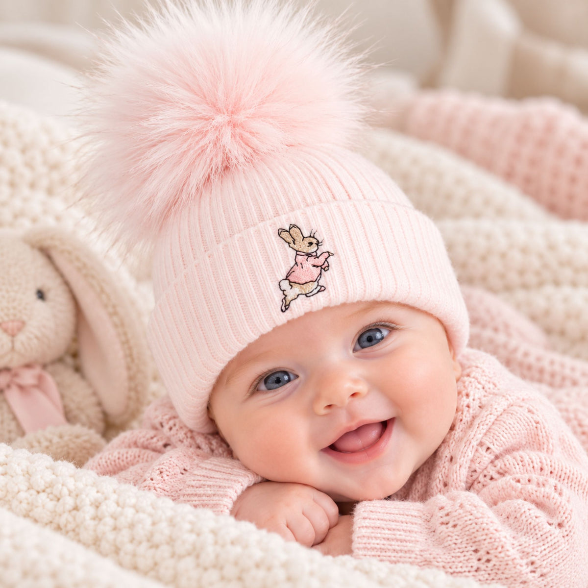 A baby wearing a light pink ribbed knit beanie with a large fluffy pom-pom and a small embroidered bunny character on the front, lying on a cream knit blanket next to a stuffed bunny.