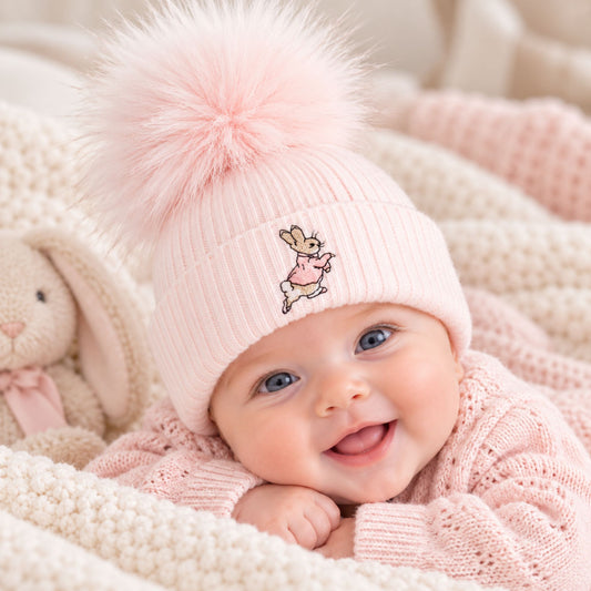 A baby wearing a light pink ribbed knit beanie with a large fluffy pom-pom and a small embroidered bunny character on the front, lying on a cream knit blanket next to a stuffed bunny.
