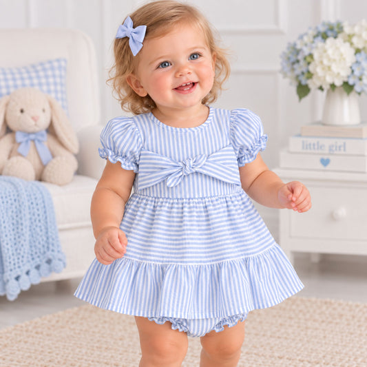A toddler girl wearing a light blue and white vertically striped baby dress with puff sleeves, a smocked bodice, and a large bow at the chest, standing in a bright nursery setting.