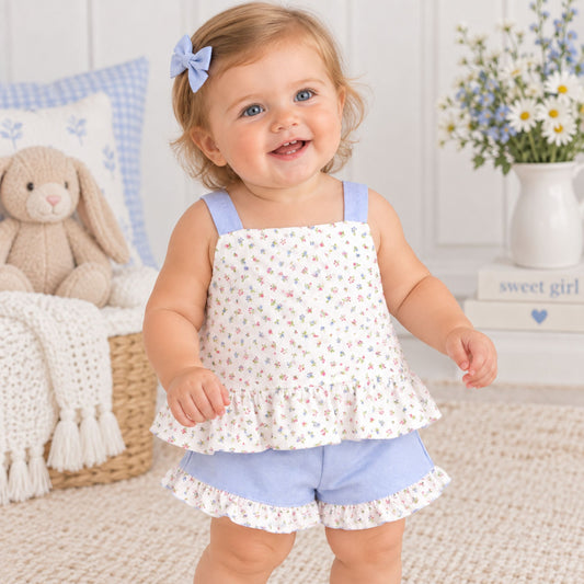 A toddler girl wearing a matching sleeveless top and shorts set with a small multicolor floral print on an off-white background and light blue trim, standing in a bright nursery setting.