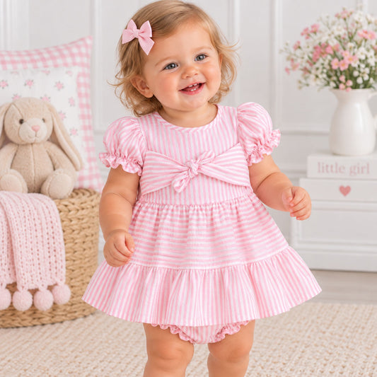 A smiling toddler girl wearing a pink and white striped tiered dress with puff sleeves and a bow on the chest, standing in a decorated nursery setting.