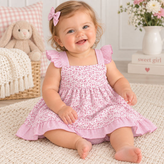 A baby girl sitting on a cream-colored rug wearing a pink and white gingham and floral print dress with ruffled shoulder straps and a matching pink bow in her hair.