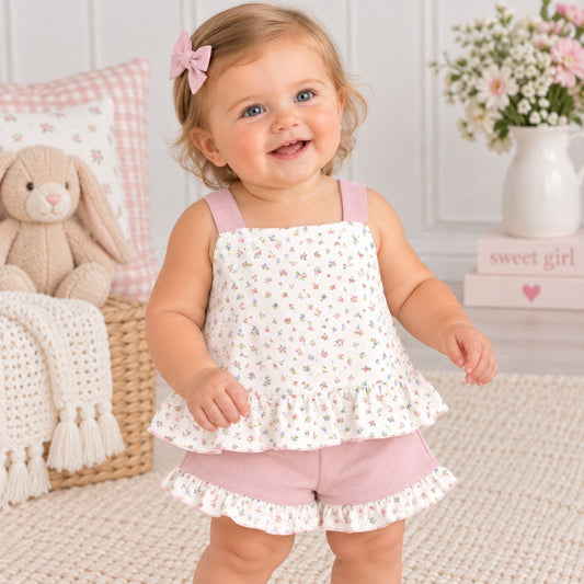 A smiling toddler girl wearing a white sleeveless romper top with a small multicolor floral print and pink ruffled trim, paired with dusty pink shorts, standing in a cozy nursery setting.