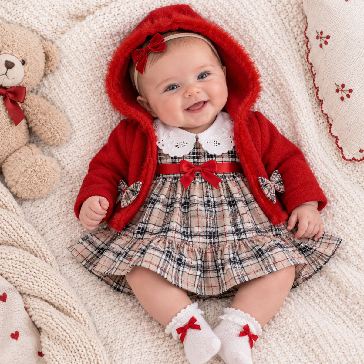 A baby wearing a red hooded cardigan and a plaid dress with white lace collar, lying on a cream knit blanket next to a teddy bear.