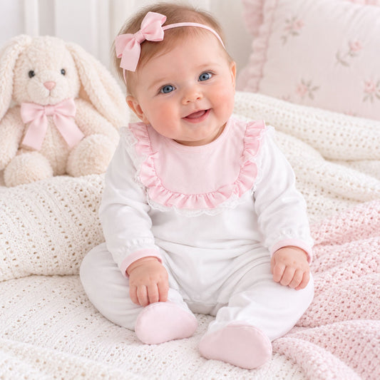A baby sitting on a soft cream knit blanket, wearing a white long-sleeve footed romper with a pink ruffled collar and matching pink booties. The baby has a pink bow headband and is posed next to a plush cream bunny toy.