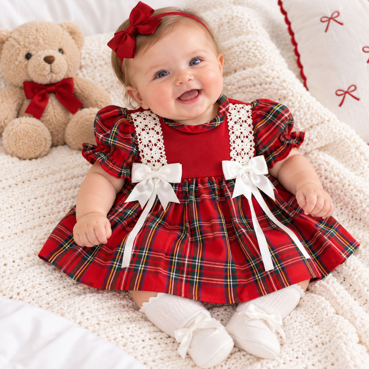 A baby wearing a red holiday plaid dress with white lace and bow details, white booties, and a red bow headband, sitting on a white knit blanket next to a teddy bear.
