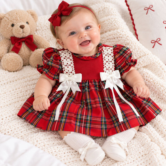 A baby wearing a red holiday plaid dress with white lace and bow details, white booties, and a red bow headband, sitting on a white knit blanket next to a teddy bear.