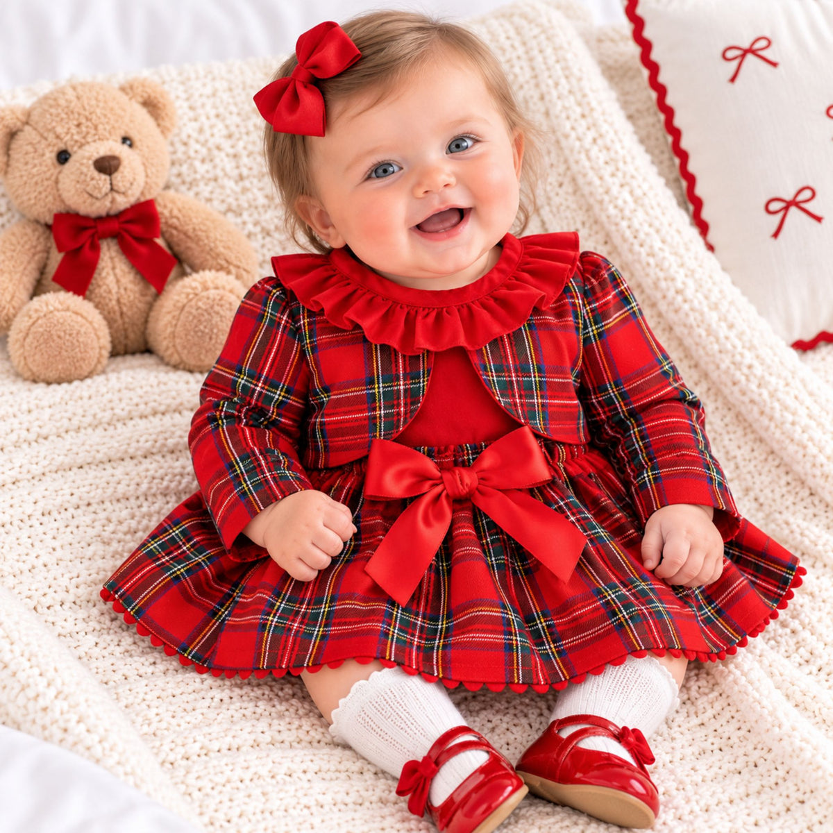 A baby girl wearing a festive red plaid dress with a large red bow at the waist, a matching red bow headband, white socks, and red shoes, sitting on a cream knit blanket next to a teddy bear.