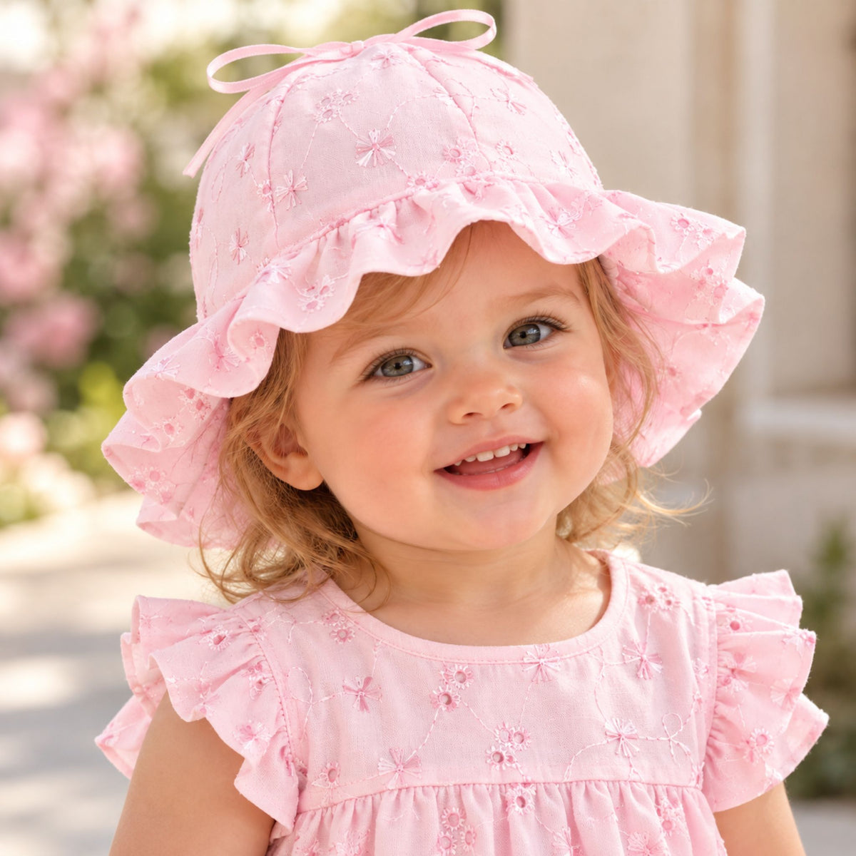 A smiling toddler girl wearing a light pink embroidered sun hat with a ruffled brim and bow, paired with a matching pink ruffle-sleeve dress, photographed outdoors against a softly blurred background.