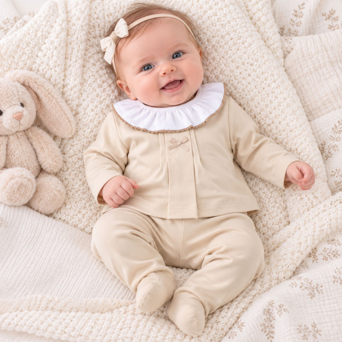 A baby lying on a textured cream blanket, wearing a beige footed romper with a white ruffled collar and a matching headband, next to a plush bunny toy.