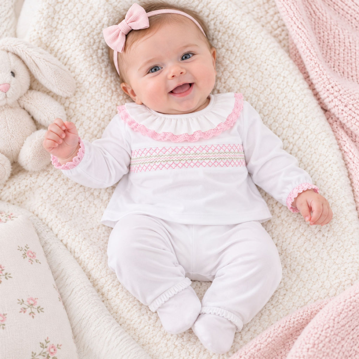 A baby wearing a white long-sleeve footed romper with pink ruffle trim and a smocked chest pattern, lying on a cream textured blanket next to a plush bunny and pink blanket.