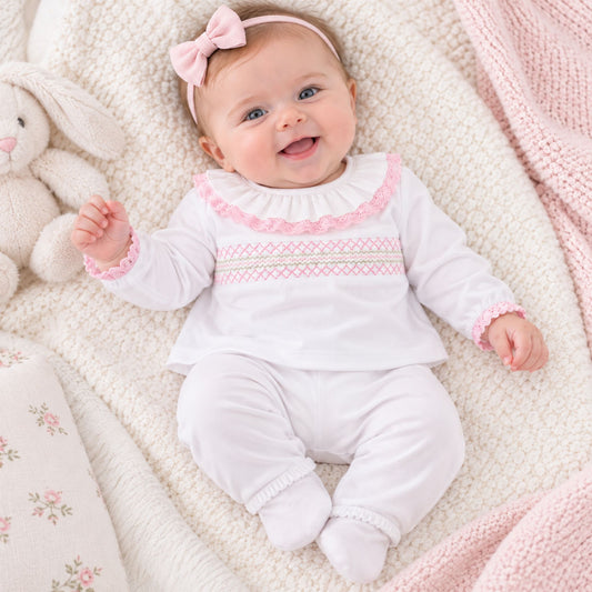 A baby wearing a white long-sleeve footed romper with pink ruffle trim and a smocked chest pattern, lying on a cream textured blanket next to a plush bunny and pink blanket.