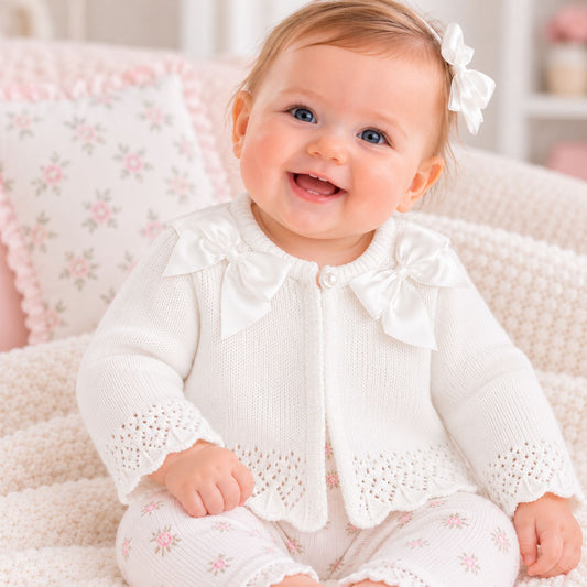 A smiling baby girl wearing a white knit cardigan with large bow details and matching white pants with pink floral prints, sitting on a soft cream blanket.