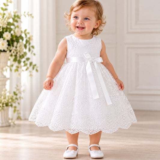 A toddler girl wearing a sleeveless white lace and tulle first communion dress with a large white satin bow at the waist, standing in a bright, elegant room.