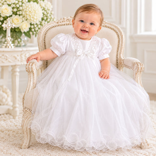 A baby girl wearing a white first communion dress sits on an ornate cream-colored armchair in a bright, elegant room. The dress features short puff sleeves, a lace-trimmed bodice with a central bow, and a full tulle skirt with ruffled trim.