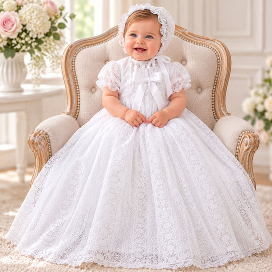 A baby girl wearing a white lace first communion dress with a matching bonnet, sitting in a classic tufted armchair with floral arrangements in the background.