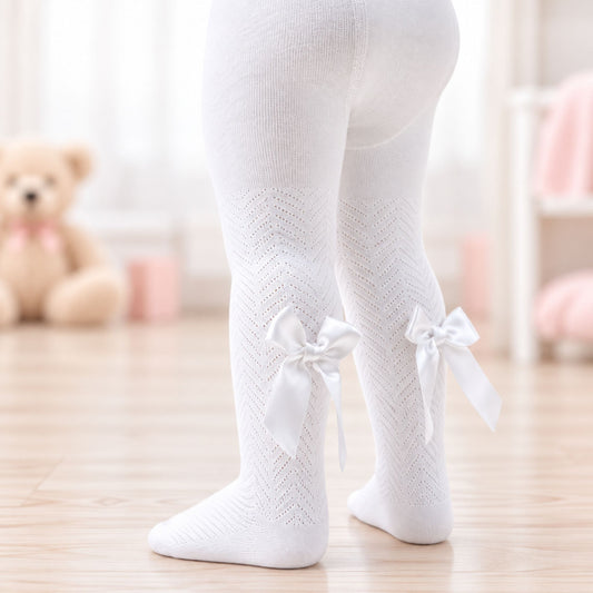 White knit tights with a textured chevron pattern and white ribbon bows at the ankles, shown on a child's legs in a softly lit room.
