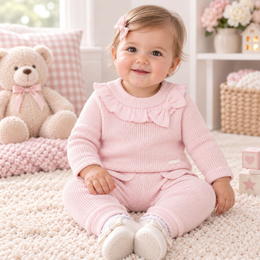 A baby girl sitting on a cream-colored textured rug in a bright, soft room. She is wearing a light pink ribbed knit two-piece outfit with a ruffled collar and bow, white shoes, and a small pink hair bow.