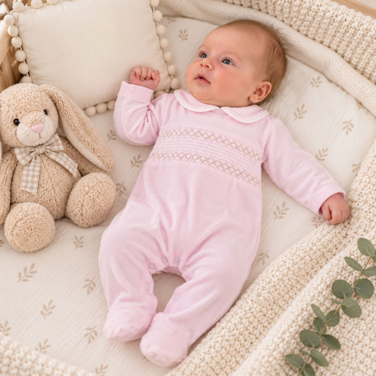 A baby lying on a cream-colored bassinet cushion, wearing a light pink footed sleeper with a decorative smocked chest panel. A beige plush bunny toy sits to the left, and a knitted blanket is visible around the bassinet.