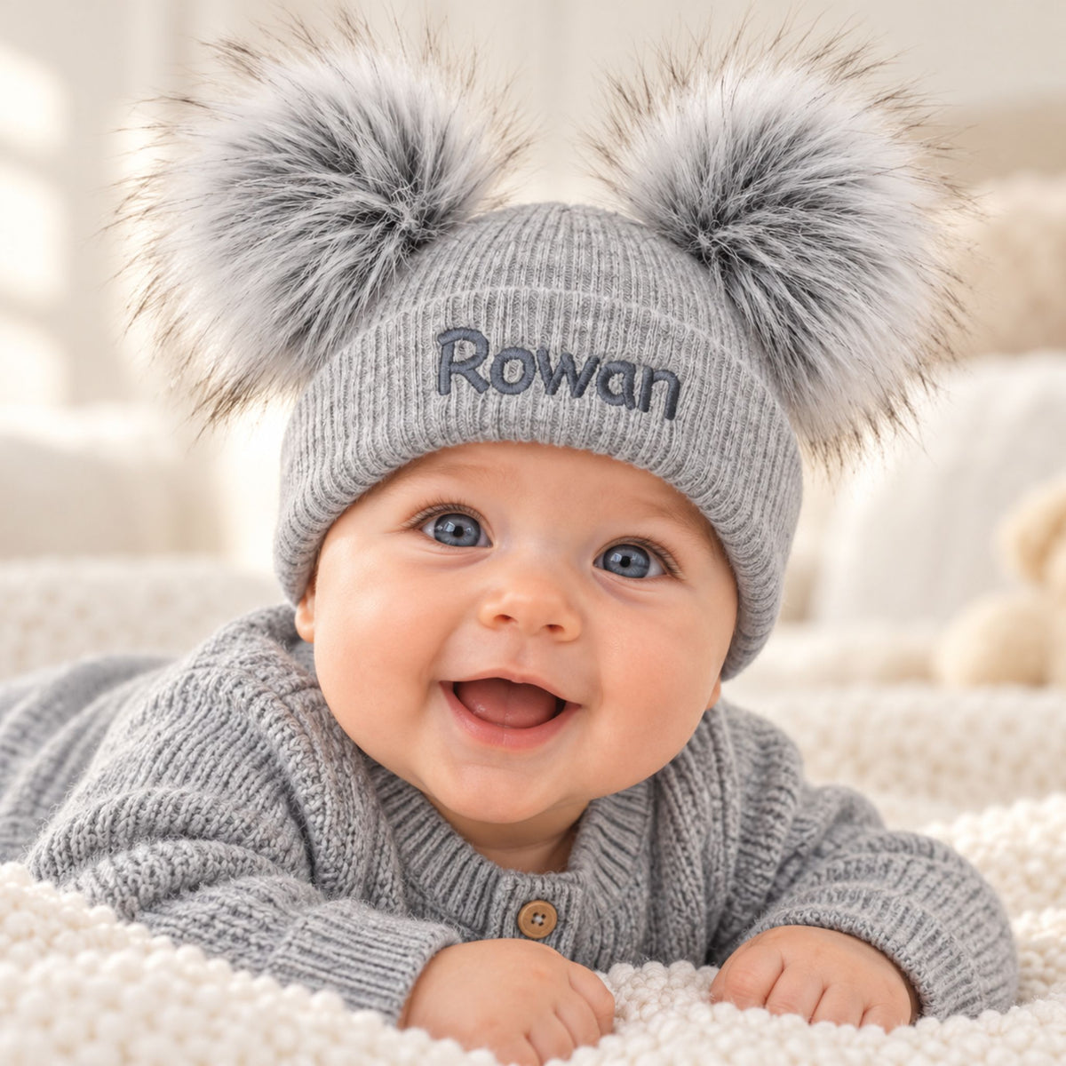 A baby lying on a soft cream blanket wearing a gray knit beanie with two large fluffy pom-poms and the name 'Rowan' embroidered on the front, along with a matching gray knit top.