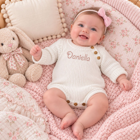 A baby lying on a pink floral blanket in a bassinet, wearing a cream ribbed long-sleeve bodysuit with 'Daniella' embroidered on the chest and pink button details. The baby is also wearing a pink bow headband and is surrounded by a plush bunny toy and pillows.