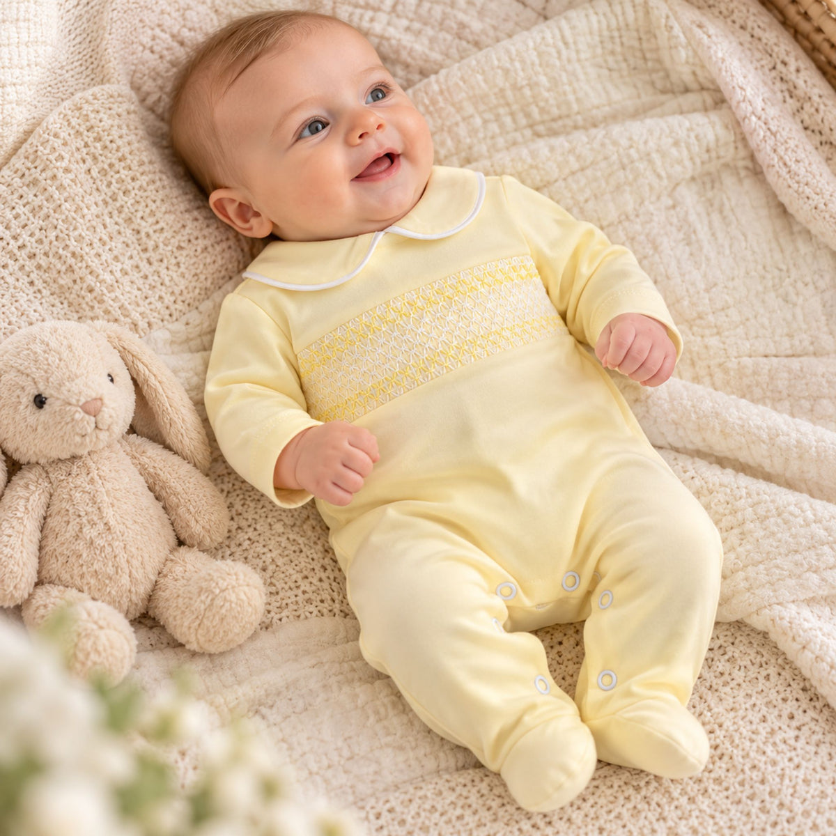 A baby lying on a cream textured blanket next to a plush bunny toy, wearing a pale yellow footed bodysuit with white trim and a decorative yoke.