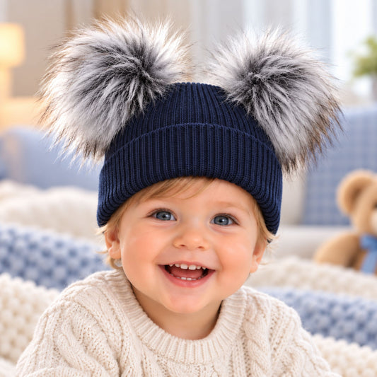 A smiling toddler wearing a navy blue ribbed knit beanie with two large fluffy pom-poms in gray, white, and black, posed in a cozy indoor setting.