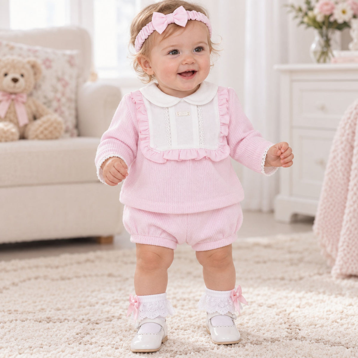 A baby girl wearing a pink ribbed long-sleeve romper with a white Peter Pan collar and ruffle trim, matching pink bubble pants, white socks with pink bows, and a pink bow headband, standing on a cream rug in a bright nursery.