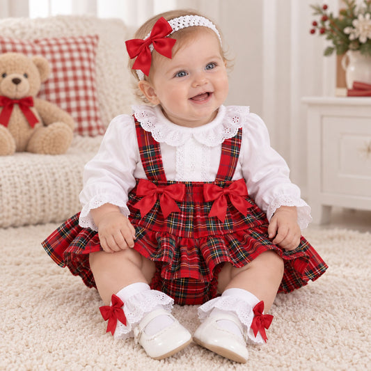 A baby girl wearing a festive holiday outfit consisting of a white long-sleeve blouse with a lace collar and a red plaid ruffled skirt with red bows, sitting on a cream carpet in a cozy room.