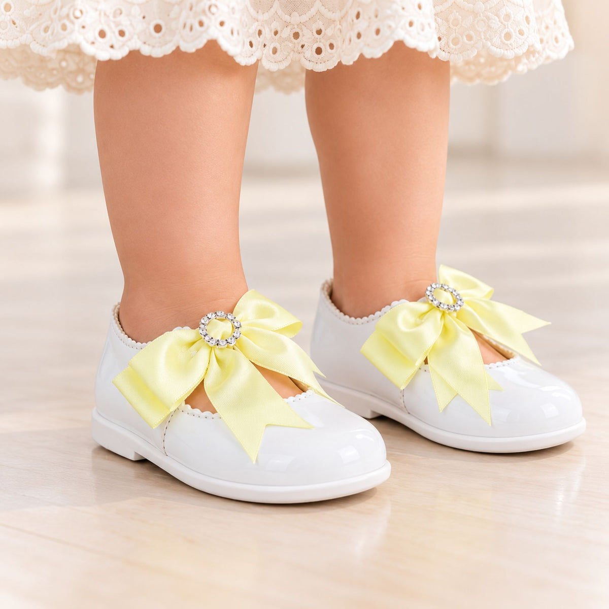 A close-up of a child's feet wearing white patent leather Mary Jane shoes with large pale yellow satin bows and crystal-embellished centers, standing on a light wood floor.