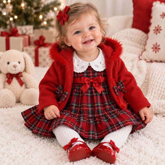 A toddler girl sitting on a white fluffy rug wearing a festive red holiday outfit with a red plaid dress, red faux-fur trimmed jacket, white lace collar, white tights, and red shoes. A teddy bear, wrapped gifts, and a decorated Christmas tree are visible in the background.