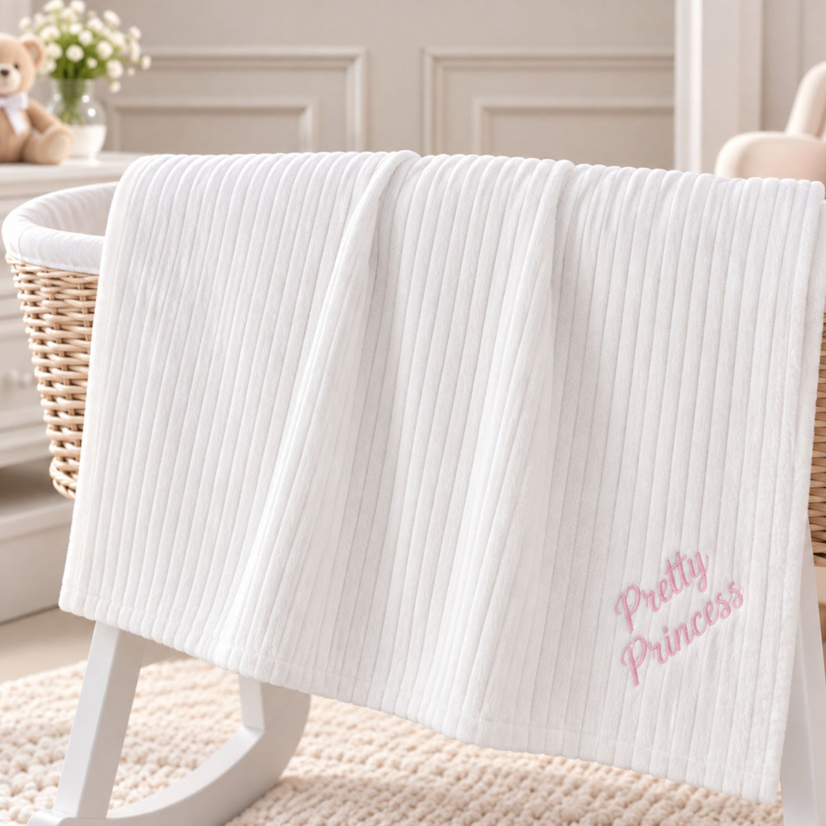A white ribbed baby blanket with pink embroidered text reading 'Pretty Princess' draped over a wicker bassinet in a softly styled nursery setting.