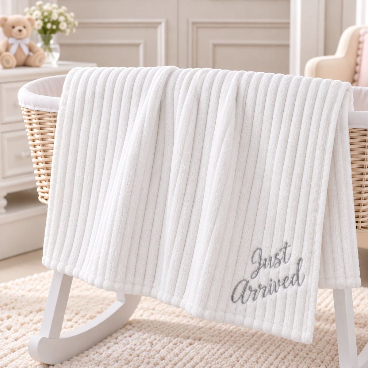 A white ribbed baby blanket with 'Just Arrived' embroidered in gray script, draped over a white wooden bassinet in a softly styled nursery setting.