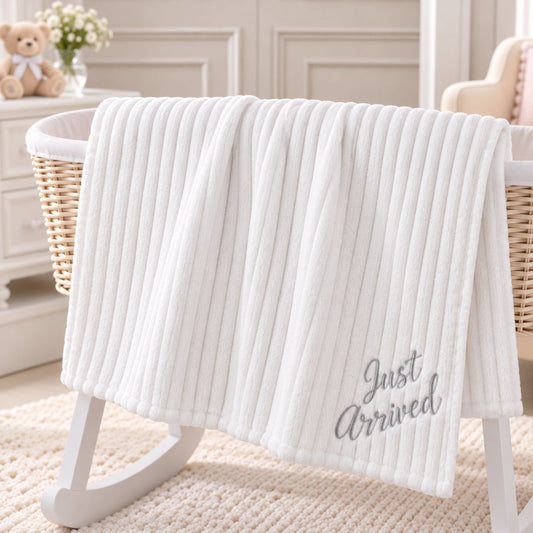 A white ribbed baby blanket with 'Just Arrived' embroidered in gray script, draped over a white wooden bassinet in a softly styled nursery setting.
