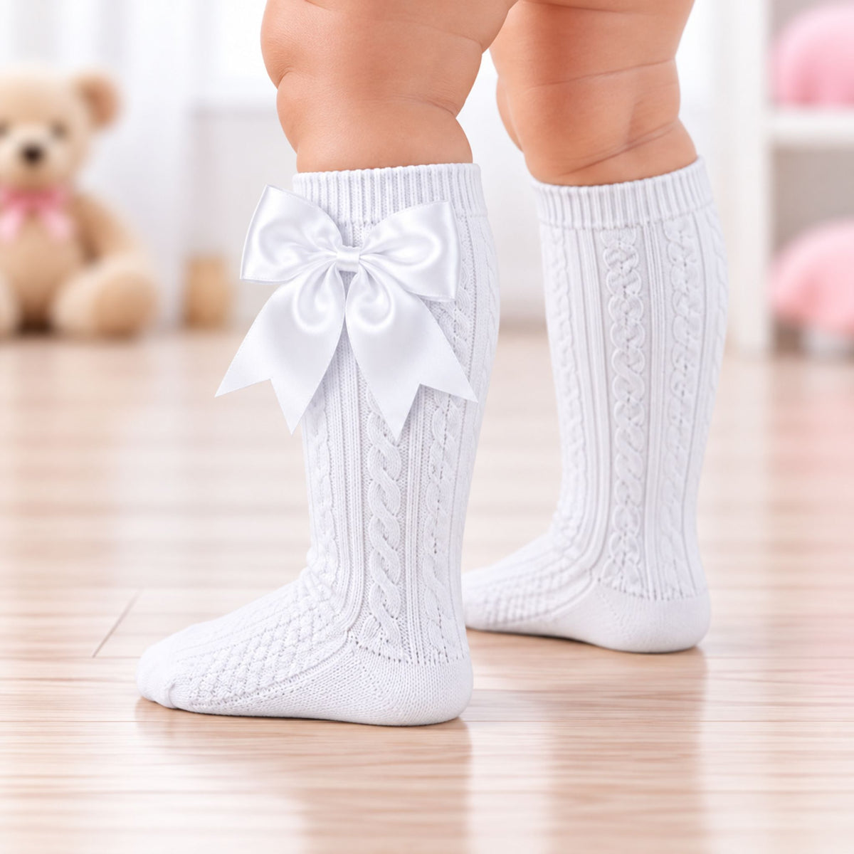 White cable-knit baby socks with a large white satin bow on the front, shown on a child's legs standing on a light wood floor.