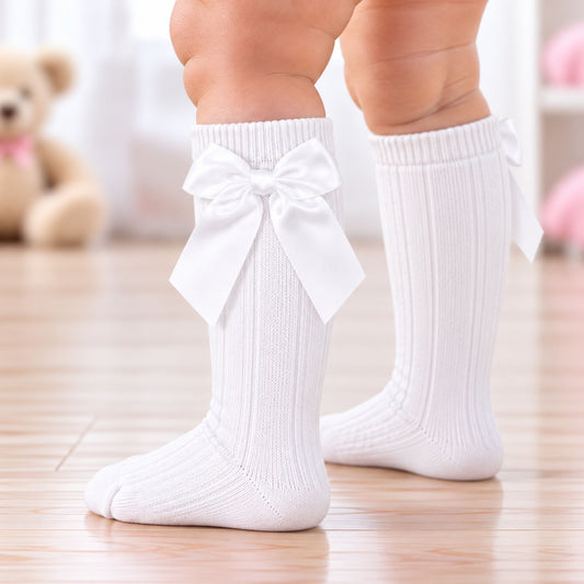 White ribbed baby knee-high socks with large bow details on the front, shown on a child's legs standing on a wooden floor.