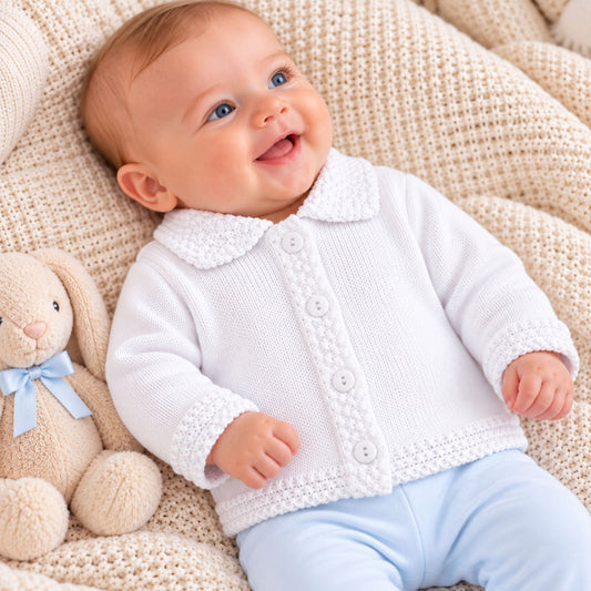 A smiling baby lying on a cream knit blanket, wearing a white knitted button-front cardigan with a textured collar and cuffs, paired with light blue pants. A beige stuffed bunny with a blue bow sits to the left.