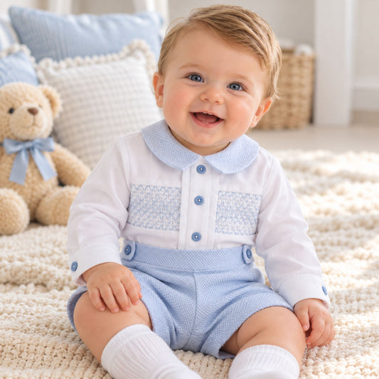 A smiling baby sitting on a cream knit rug, wearing a white long-sleeve top with a light blue patterned chest panel and collar, light blue shorts, and white socks. A teddy bear and pillows are visible in the softly lit background.