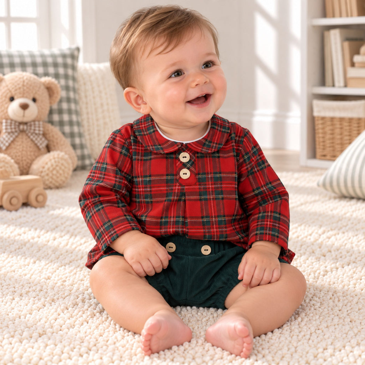 A baby sitting on a white textured rug in a bright room, wearing a red and green plaid long-sleeve shirt with three wooden buttons and dark green shorts.