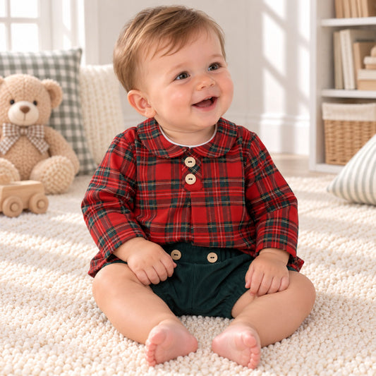 A baby sitting on a white textured rug in a bright room, wearing a red and green plaid long-sleeve shirt with three wooden buttons and dark green shorts.