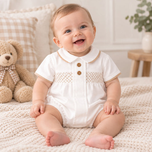A smiling baby sitting on a cream knit blanket in a nursery, wearing a white short-sleeve romper with a beige Peter Pan collar, embroidered chest details, and two brown buttons.