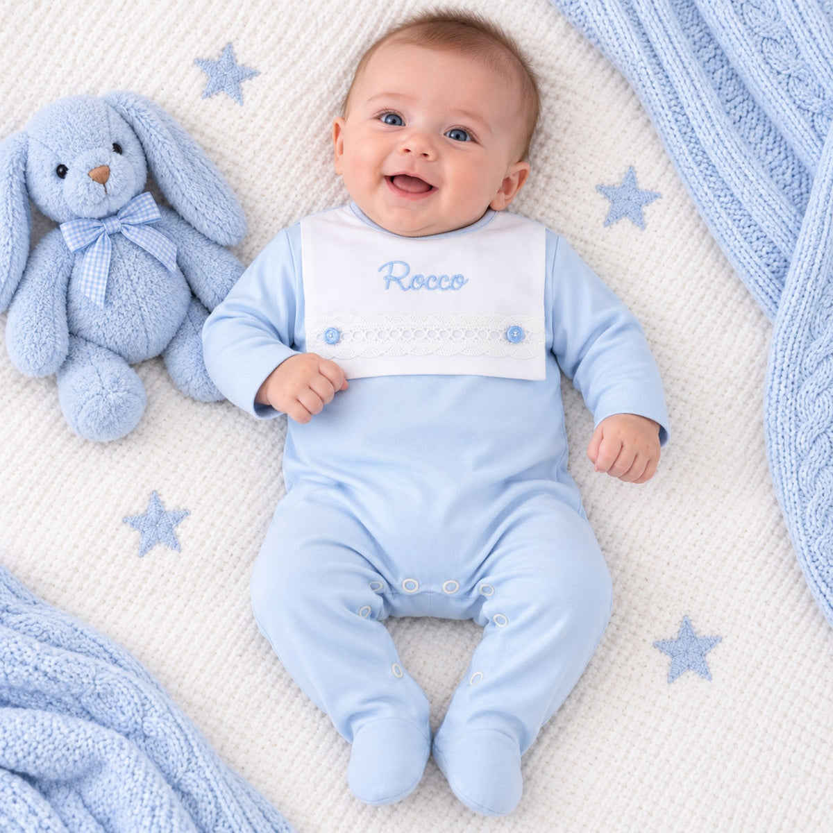A smiling baby wearing a light blue footed sleepsuit with a white bib embroidered with the name 'Rocco' lies on a white textured blanket with blue star prints, next to a plush blue bunny toy.