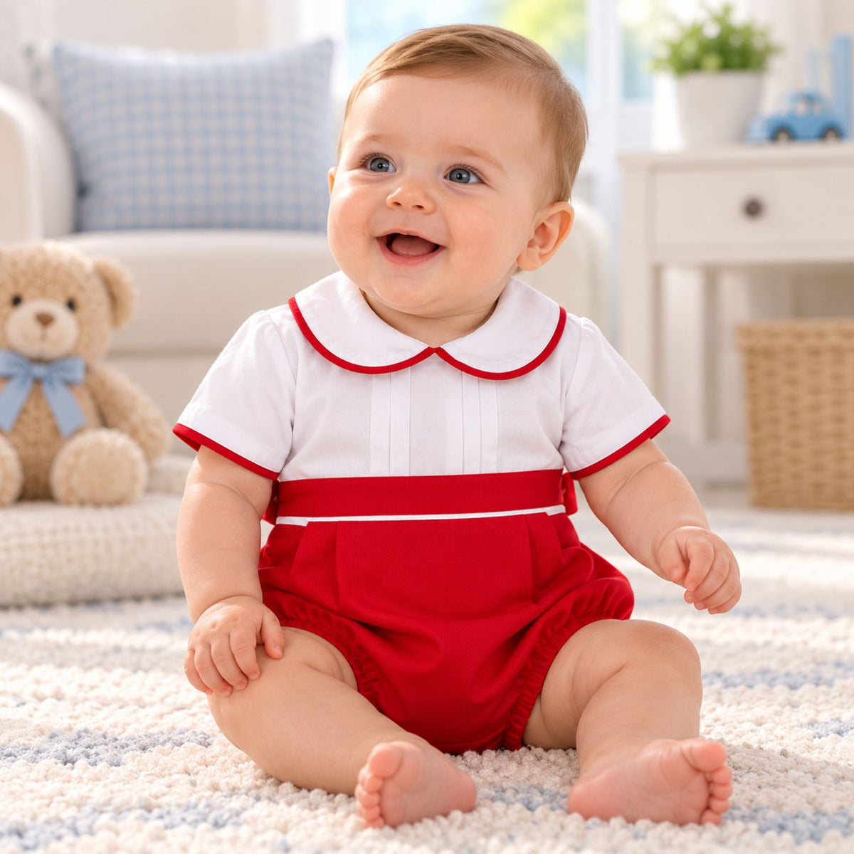 A smiling baby sitting on a white textured rug in a bright nursery, wearing a short-sleeve romper with a white top featuring a red Peter Pan collar and red trim, paired with red bloomers.