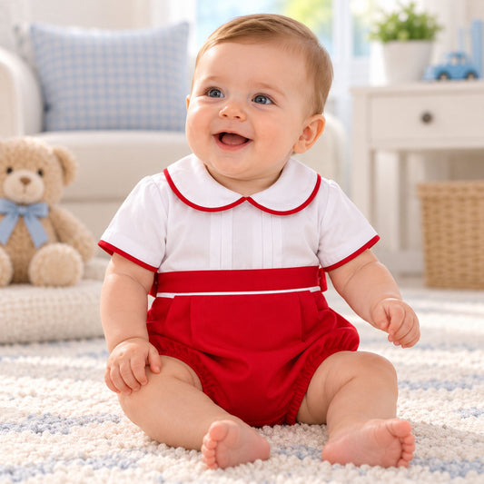 A smiling baby sitting on a white textured rug in a bright nursery, wearing a short-sleeve romper with a white top featuring a red Peter Pan collar and red trim, paired with red bloomers.
