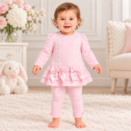 A toddler girl stands barefoot on a white shag rug wearing a light pink long-sleeve dress with ruffled trim, decorative vertical details, and large bows at the waist, paired with matching pink leggings. The background shows a softly styled nursery interior with a white bunny plush, flowers, and a chair.